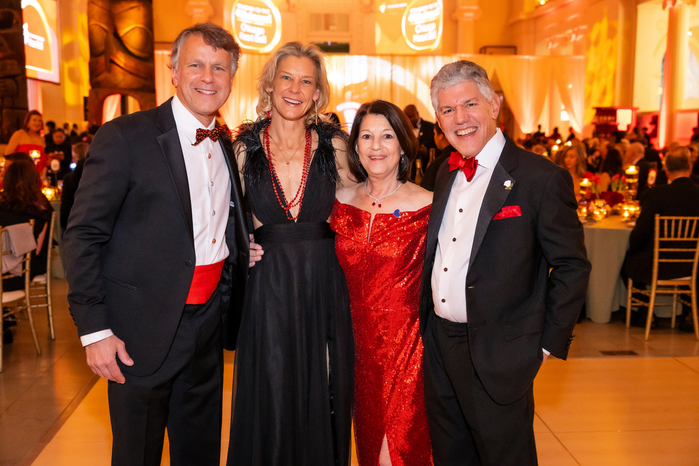 Four adults in formal attire posing and smiling at a gala event. Two men in tuxedos with red accents and two women in red and black dresses stand together.