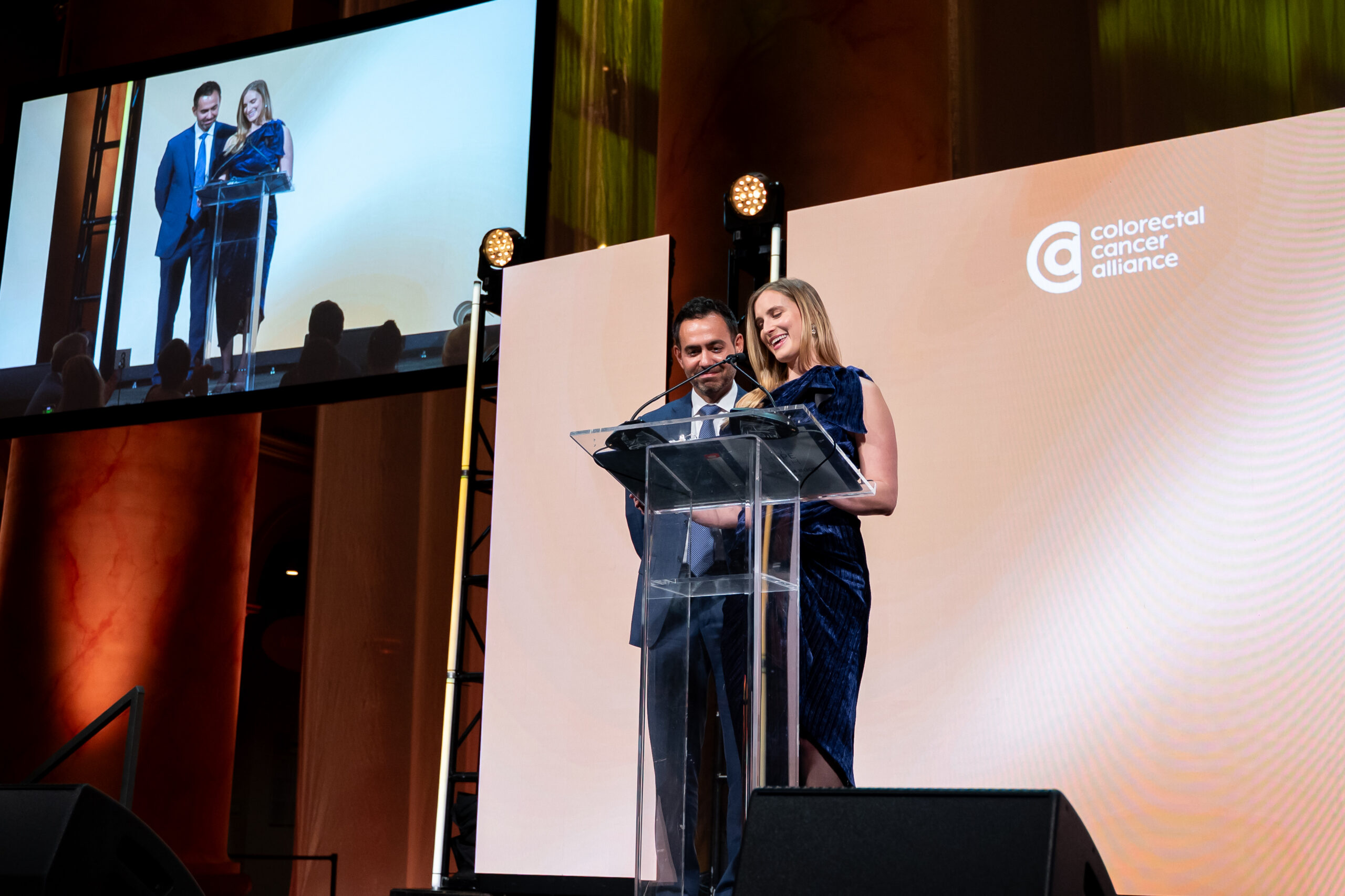 Two presenters stand at a clear podium on stage during a colorectal cancer alliance event, with a large screen behind them showing the duo; the woman wears a blue dress and the man a suit.