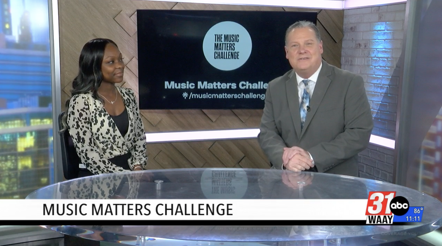 News anchors sit at a glass desk in a studio, with a screen reading 'Music Matters Challenge' behind them.